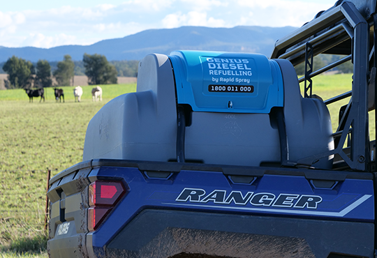 A blue and gray Centius Diesel Refueling tank mounted on the back of a blue and gray Ranger utility vehicle, parked in a grassy field with cows and mountains in the background.